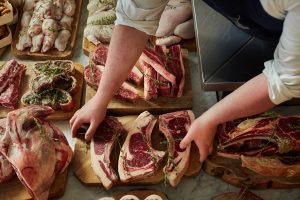A butcher in a white shirt and dark apron arranges large cuts of beef on a wooden board, on a table with a varied assortment of meats presented in a similar way.